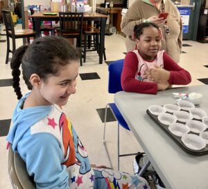 A girl in a blue sweatshirt and a girl in a red shirt sitting at a table and smiling as they are making muffins.