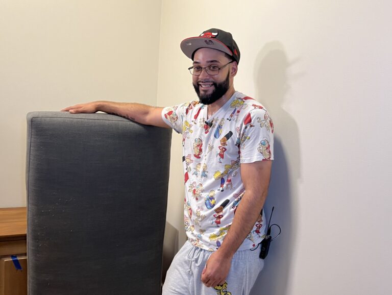 A young man in a hat and patterned shirt and light jeans resting his hand on a couch cushion