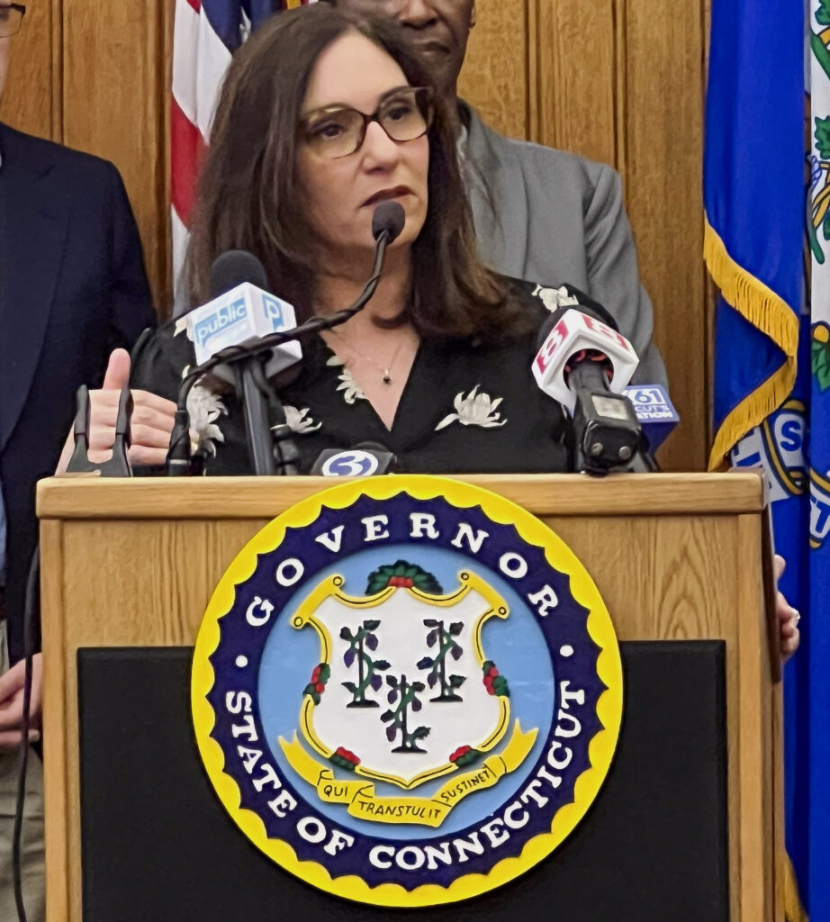 CEO Kara Capone speaking to a group of people at the Governor's podium, with the seal of the State of Connecticut over a black box.